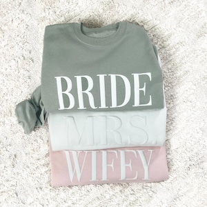 May include: Three sweatshirts stacked on top of each other. The top sweatshirt is sage green with the word "BRIDE" in white lettering. The middle sweatshirt is white with the word "MRS." in white lettering. The bottom sweatshirt is pink with the word "WIFEY" in white lettering.