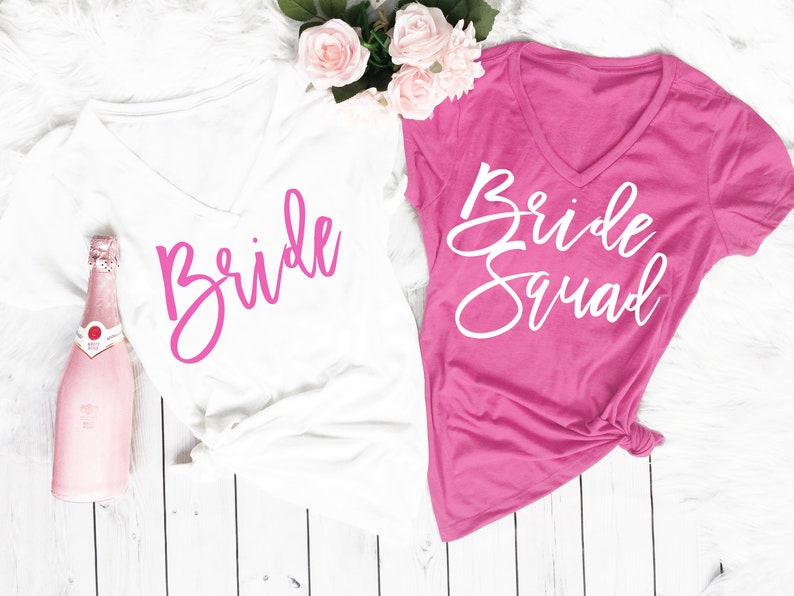Puede incluir: Dos camisetas blancas de cuello en V con letras rosas. Una camiseta dice "Bride" y la otra dice "Bride Squad".