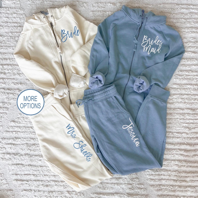May include: Two pairs of matching sweatpants and hoodies. One set is light blue with the text "Bride Maid" on the hoodie and "Jessica" on the sweatpants. The other set is cream with the text "Bride" on the hoodie and "Mrs. Shields" on the sweatpants.