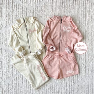 May include: Two matching sets of cream and pink sweatshirts and sweatpants. The cream set has the text "Bride" embroidered on the sweatshirt and "Mrs. Miller" embroidered on the sweatpants. The pink set has the text "Bride's Maid" embroidered on the sweatshirt and "Ashley" embroidered on the sweatpants.
