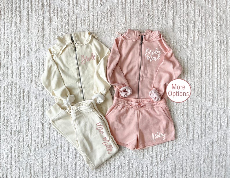 May include: Two matching sets of cream and pink sweatshirts and sweatpants. The cream set has the text "Bride" embroidered on the sweatshirt and "Mrs. Miller" embroidered on the sweatpants. The pink set has the text "Bride's Maid" embroidered on the sweatshirt and "Ashley" embroidered on the sweatpants.