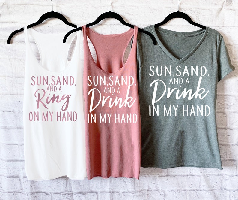 May include: Three tank tops with the text "Sun, Sand, and a Ring on my hand", "Sun, Sand, and a Drink in my hand", and "Sun, Sand, and a Drink in my hand" printed on them. The tank tops are white, pink, and gray.