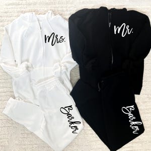 May include: A white and black matching set of zippered hoodies and sweatpants. The white hoodie has "Mrs." printed on the front and "Barker" printed on the leg of the sweatpants. The black hoodie has "Mr." printed on the front and "Barker" printed on the leg of the sweatpants.
