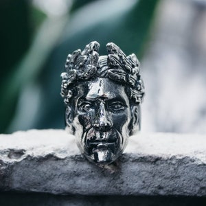May include: A silver ring with a detailed sculpture of a face wearing a crown of leaves.