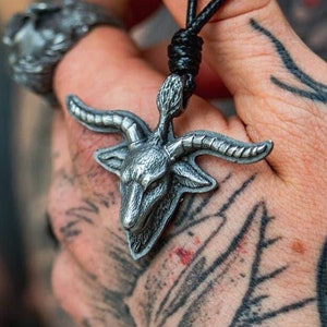 May include: A silver pendant necklace featuring a detailed goat head with horns. The pendant is hanging from a black cord.