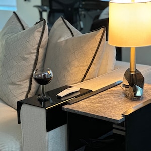 May include: A modern living room scene featuring a sofa with patterned pillows and a black side table. A glass of red wine sits on a black tray attached to the sofa. A lamp with a geometric base and a marble-look top is on the side table.