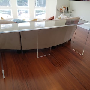 May include: A clear acrylic console table with a rectangular top and four legs. The table is in a living room setting with a couch in the background.