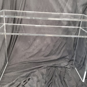 May include: A clear acrylic console table with two shelves. The table is made of thick, clear acrylic and has a modern, minimalist design.