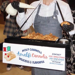 May include: A person is shown preparing cannoli pastries. The pastries are in a black tray with compartments. The tray has a sign that reads "Holy Cannoli Amalfi Cannolis Delicious & Flavorful". The person is wearing a white shirt and a gray apron.