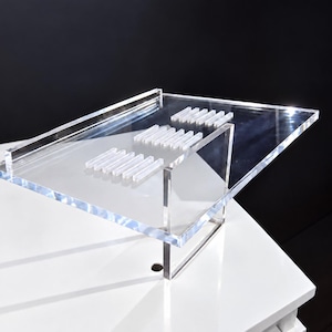 May include: A clear acrylic slanted display stand featuring a rectangular surface with three sets of raised, parallel rectangular bars arranged in a row. The stand is constructed from transparent material with a sturdy vertical support base.