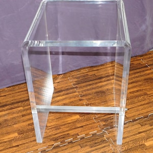 May include: A clear acrylic bar stool with a rectangular seat and a footrest. The stool has a modern design with a transparent appearance, showcasing the surrounding environment. The legs are straight and the footrest is a horizontal bar.