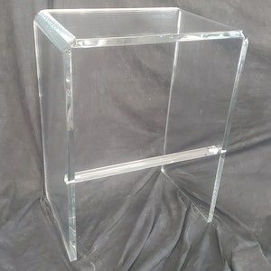 Könnte beinhalten: Ein Barhocker aus klarem Acryl mit einem U-förmigen Rahmen, abgerundeten oberen Ecken und einer horizontalen Fußstütze, die die beiden Seitenteile verbindet. Der Hocker besteht aus transparentem Material mit glatten, polierten Kanten.