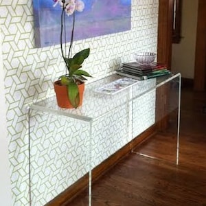 May include: A clear acrylic console table with a white and gold geometric patterned wallpaper behind it. A potted orchid with pink and white flowers sits on the table.