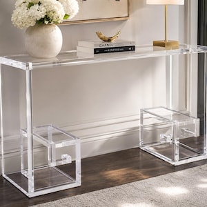 May include: A rectangular console table constructed from clear acrylic. The table features a flat top surface supported by two geometric, open-frame bases on each side, each consisting of interconnected square and rectangular acrylic panels.