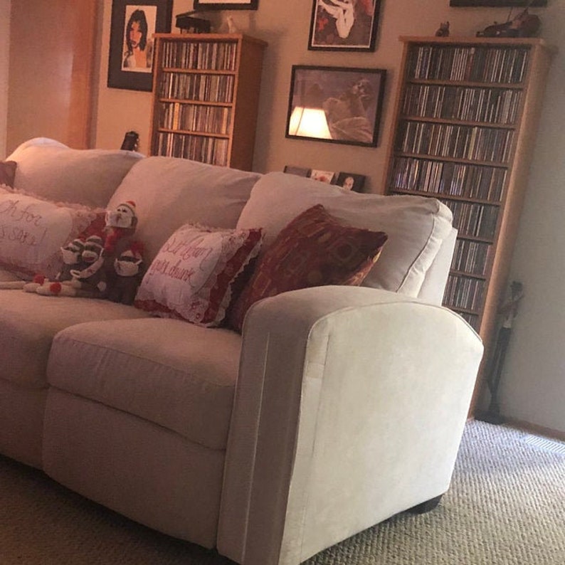 Puede incluir: Un sof&aacute; tapizado en color beige claro con varios cojines decorativos, incluido uno con borde rojo y texto bordado, junto a un peluche de mono hecho de calcetines. El sof&aacute; est&aacute; situado en una habitaci&oacute;n con dos torres de madera para discos.