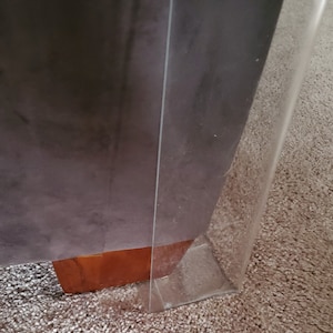 Puede incluir: Un protector de pata de mueble de acr&iacute;lico transparente colocado alrededor de una esquina de mueble tapizada en gris oscuro con una base de pata de madera marr&oacute;n claro visible. El protector transparente crea una barrera.