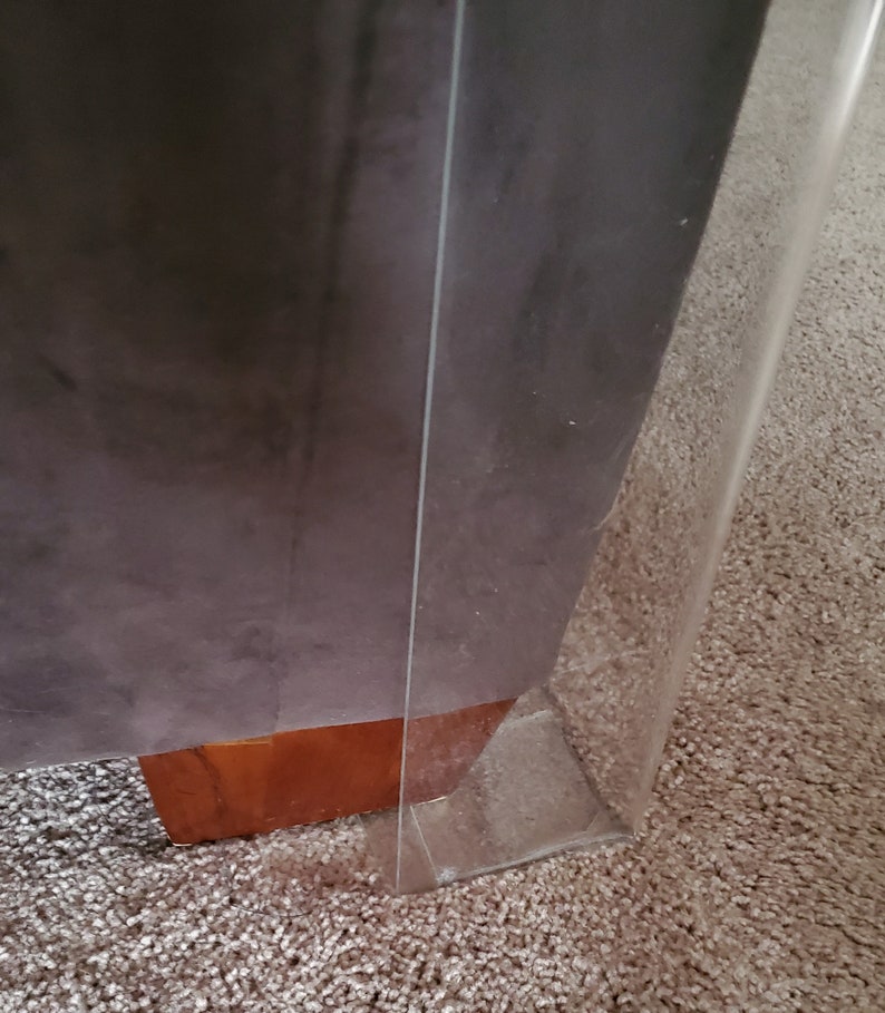 Puede incluir: Un protector de pata de mueble de acr&iacute;lico transparente colocado alrededor de una esquina de mueble tapizada en gris oscuro con una base de pata de madera marr&oacute;n claro visible. El protector transparente crea una barrera.