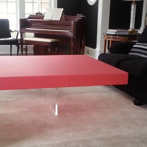 May include: A square coffee table with a solid, smooth, matte red tabletop surface. The table is supported by a clear, transparent acrylic base consisting of two intersecting vertical panels that form a cross shape beneath the center.