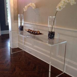 May include: A clear acrylic console table with a white base and a white wall behind it. Two clear glass vases with white orchids are on the table. A decorative bowl with gold-colored orbs is on the table.
