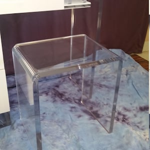 May include: A clear acrylic side table with a square top and curved legs. The table is sitting on a blue and white patterned surface.