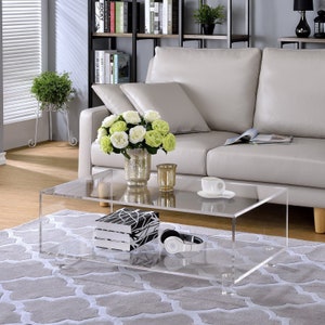 May include: A clear acrylic coffee table with a lower shelf. The table has a modern design and is perfect for a living room or den. The table is shown with a vase of flowers, a book, and a pair of headphones.