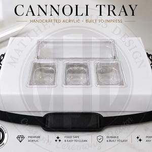 May include: A white rectangular acrylic cannoli tray featuring one long top compartment and three smaller bottom compartments. The tray includes a detachable black nylon shoulder strap with a padded section and metal clips on both sides.