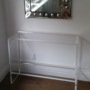 May include: A clear acrylic console table featuring a rectangular top surface and a lower shelf, constructed from a single continuous piece of transparent material with rounded edges, providing a sleek and open appearance for interior spaces.