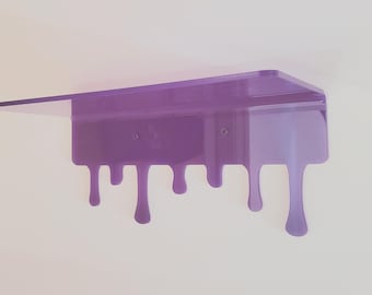 May include: A purple acrylic wall shelf featuring a flat horizontal surface and a vertical back panel with a decorative dripping liquid design along the bottom edge. The shelf includes two visible circular mounting holes for installation.