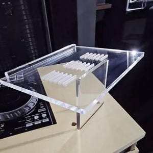 May include: A clear acrylic DJ equipment stand with a multi-tiered design. The stand features a rectangular top with several small, white rectangular supports. The stand is placed on a light beige surface, with a black DJ mixer visible in the background.