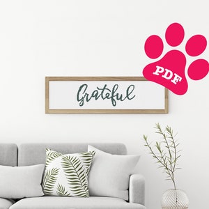 Peut inclure: Un canapé blanc avec un coussin à motif de feuilles vertes, une table basse blanche avec une pile de livres et une tasse blanche, et une petite table d'appoint en bois avec un vase en verre transparent avec des branches vertes. Une pancarte encadrée avec le mot "grateful" en écriture noire est accrochée au mur au-dessus du canapé. Une empreinte de patte rose avec le mot "PDF" en texte blanc est dans le coin supérieur droit de l'image.