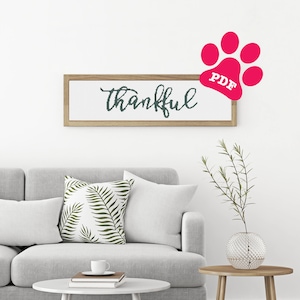 Può includere: Un divano grigio con un cuscino bianco con un motivo a foglie verdi. Un tavolino da caffè bianco con due libri e una tazza bianca in cima. Un piccolo tavolino da salotto in legno con un vaso di vetro trasparente con una pianta verde. Un cartello incorniciato con la parola "grato" in scrittura nera. Un'impronta di zampa rosa con la parola "PDF" in testo bianco.