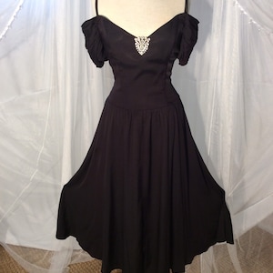 May include: A black off-the-shoulder dress with a fitted bodice and a flowing skirt. The dress has a jeweled embellishment at the neckline.