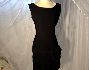 May include: A black sleeveless dress with a tiered skirt and fringe trim. The dress is on a white mannequin against a white sheer background.