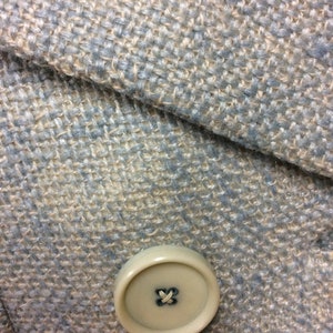 May include: A close-up of a light blue and beige tweed fabric with a single beige button. The button has a simple cross stitch in the center.