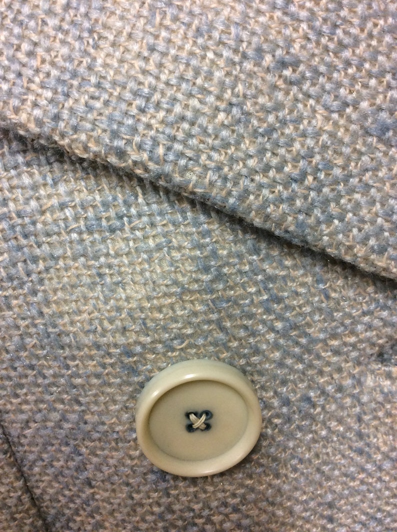 May include: A close-up of a light blue and beige tweed fabric with a single beige button. The button has a simple cross stitch in the center.