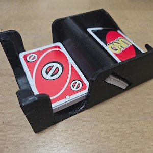 May include: A black plastic card holder with two compartments. One compartment holds a deck of red and white Uno cards, and the other holds a few cards with the word UNO on them. The holder is on a wooden surface.
