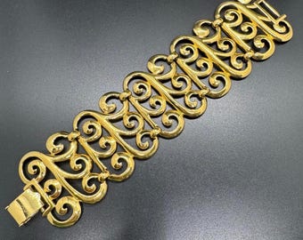 May include: A gold-toned bracelet with an ornate, openwork design. The bracelet features a series of interconnected, scroll-like patterns. The clasp is a simple, rectangular design. The jewelry is displayed on a dark surface.