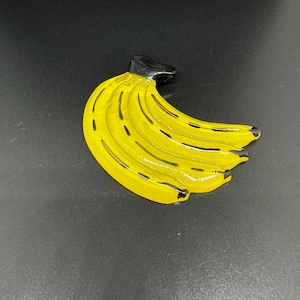 Puede incluir: Un broche con forma de racimo de plátanos de color amarillo brillante con un acabado lustroso y un tallo negro. El diseño muestra un grupo de cinco plátanos curvados con líneas negras que acentúan las formas y una superficie lisa.