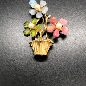Puede incluir: Broche floral dorado con tres racimos de flores en un jarr&oacute;n. Las flores est&aacute;n hechas de piedras azules, verdes y rosas, cada una con un centro de perla. El broche tiene una base de jarr&oacute;n detallada y texturizada.