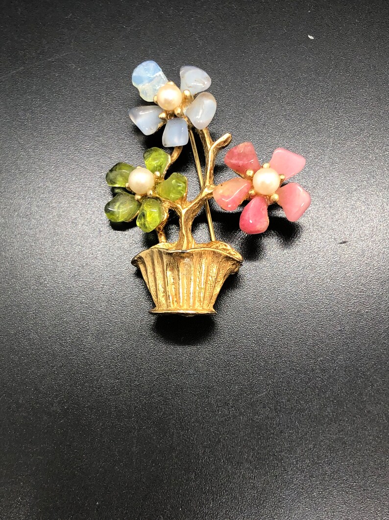 Puede incluir: Broche floral dorado con tres racimos de flores en un jarr&oacute;n. Las flores est&aacute;n hechas de piedras azules, verdes y rosas, cada una con un centro de perla. El broche tiene una base de jarr&oacute;n detallada y texturizada.