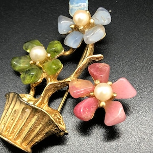 Puede incluir: Broche floral dorado con tres grupos de flores con p&eacute;talos de piedras preciosas en tonos rosa, verde y azul. Cada flor tiene un centro de perla. El broche est&aacute; dise&ntilde;ado con un jarr&oacute;n dorado.