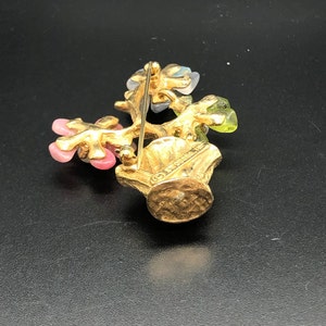 Puede incluir: Broche dorado con una cesta rebosante de flores coloridas. Las flores son rosas, verdes y azules, con un acabado dorado texturizado en la cesta y los tallos. El broche est&aacute; dise&ntilde;ado para ser usado en la ropa.