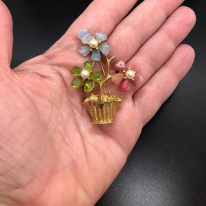 Puede incluir: Broche floral dorado con tres flores en una cesta. Las flores est&aacute;n hechas de piedras azules, verdes y rosas, cada una con un centro de perla. El broche se sostiene en una mano sobre un fondo negro.