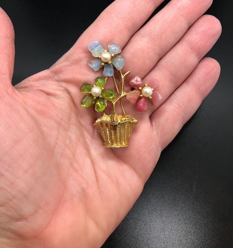 Puede incluir: Broche floral dorado con tres flores en una cesta. Las flores est&aacute;n hechas de piedras azules, verdes y rosas, cada una con un centro de perla. El broche se sostiene en una mano sobre un fondo negro.