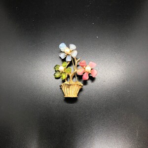 Puede incluir: Broche de metal dorado con un arreglo floral en una cesta. Las flores est&aacute;n hechas de piedras transl&uacute;cidas de colores en tonos azules, verdes y rosas, cada una con un centro de perla. El broche est&aacute; sobre un fondo negro.