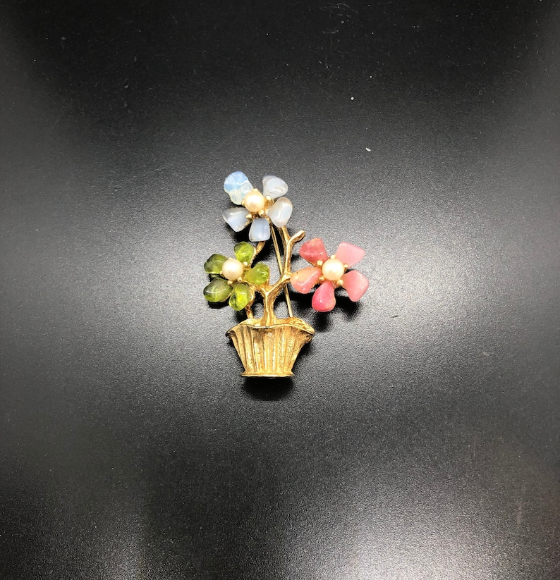Puede incluir: Broche de metal dorado con un arreglo floral en una cesta. Las flores est&aacute;n hechas de piedras transl&uacute;cidas de colores en tonos azules, verdes y rosas, cada una con un centro de perla. El broche est&aacute; sobre un fondo negro.
