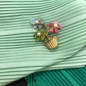 Puede incluir: Broche floral dorado con tres flores con p&eacute;talos de piedras preciosas de colores y un centro de perla, en un jarr&oacute;n dorado. El broche est&aacute; sobre una tela plisada verde claro.