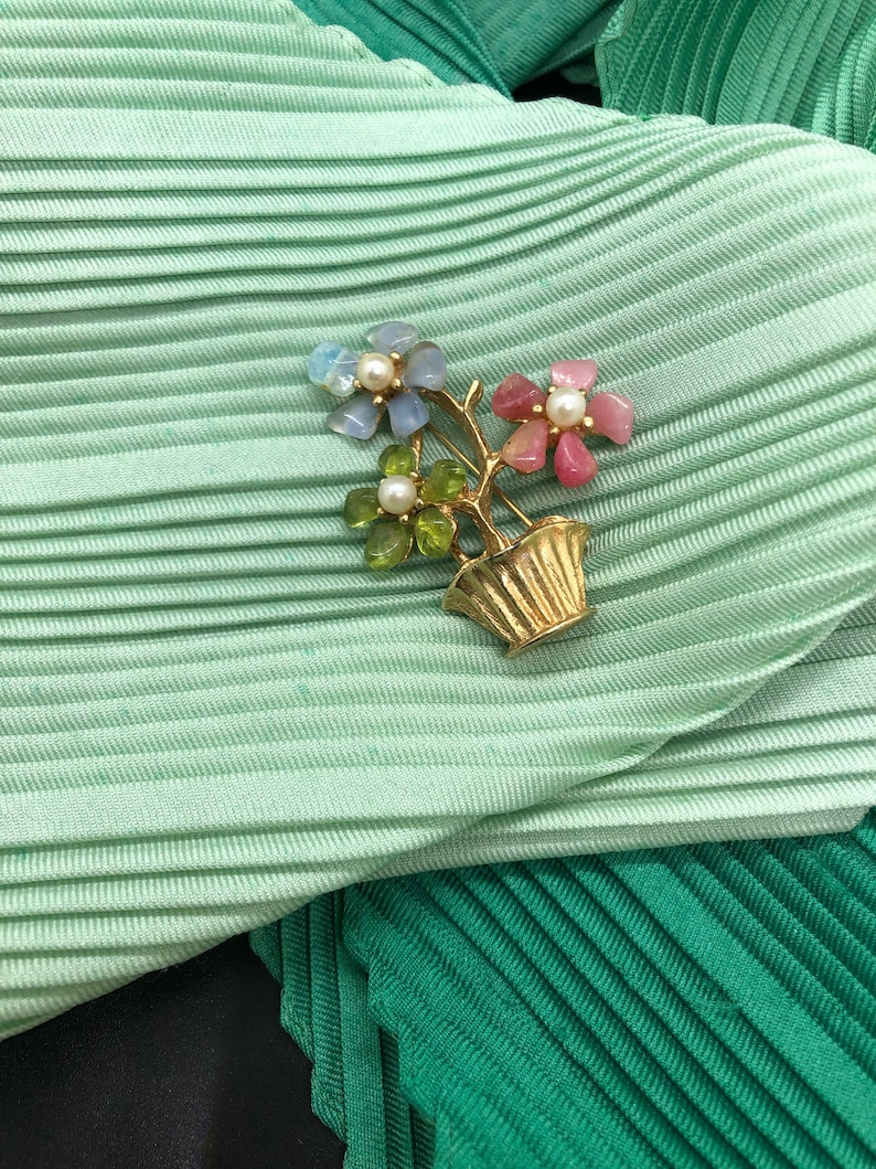 Puede incluir: Broche floral dorado con tres flores con p&eacute;talos de piedras preciosas de colores y un centro de perla, en un jarr&oacute;n dorado. El broche est&aacute; sobre una tela plisada verde claro.