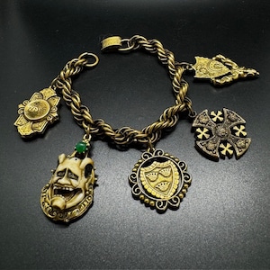 Puede incluir: Un brazalete de oro con cadena trenzada y cinco dijes. Los dijes incluyen un escudo con tres coronas, una cruz con un diseño central, una pequeña piedra verde, una máscara con una sonrisa de dientes y un diseño decorativo con una flor de lis.
