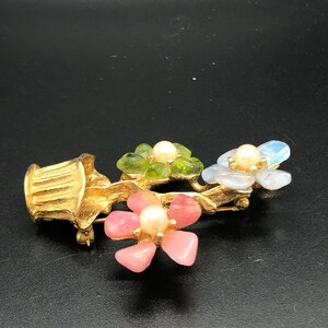 Puede incluir: Broche floral dorado con tres grupos de flores, cada uno con un centro de perla y p&eacute;talos de piedra de colores. El broche tiene un acabado dorado texturizado y un cierre de alfiler. Las flores son rosas, verdes y azules.